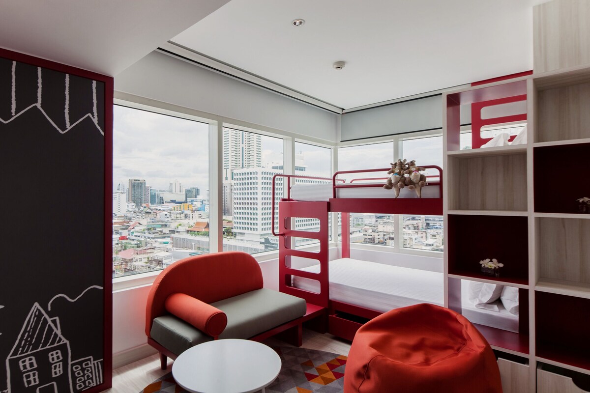 Habitación moderna con vista a la ciudad, literas, una silla roja y una mesa pequeña.