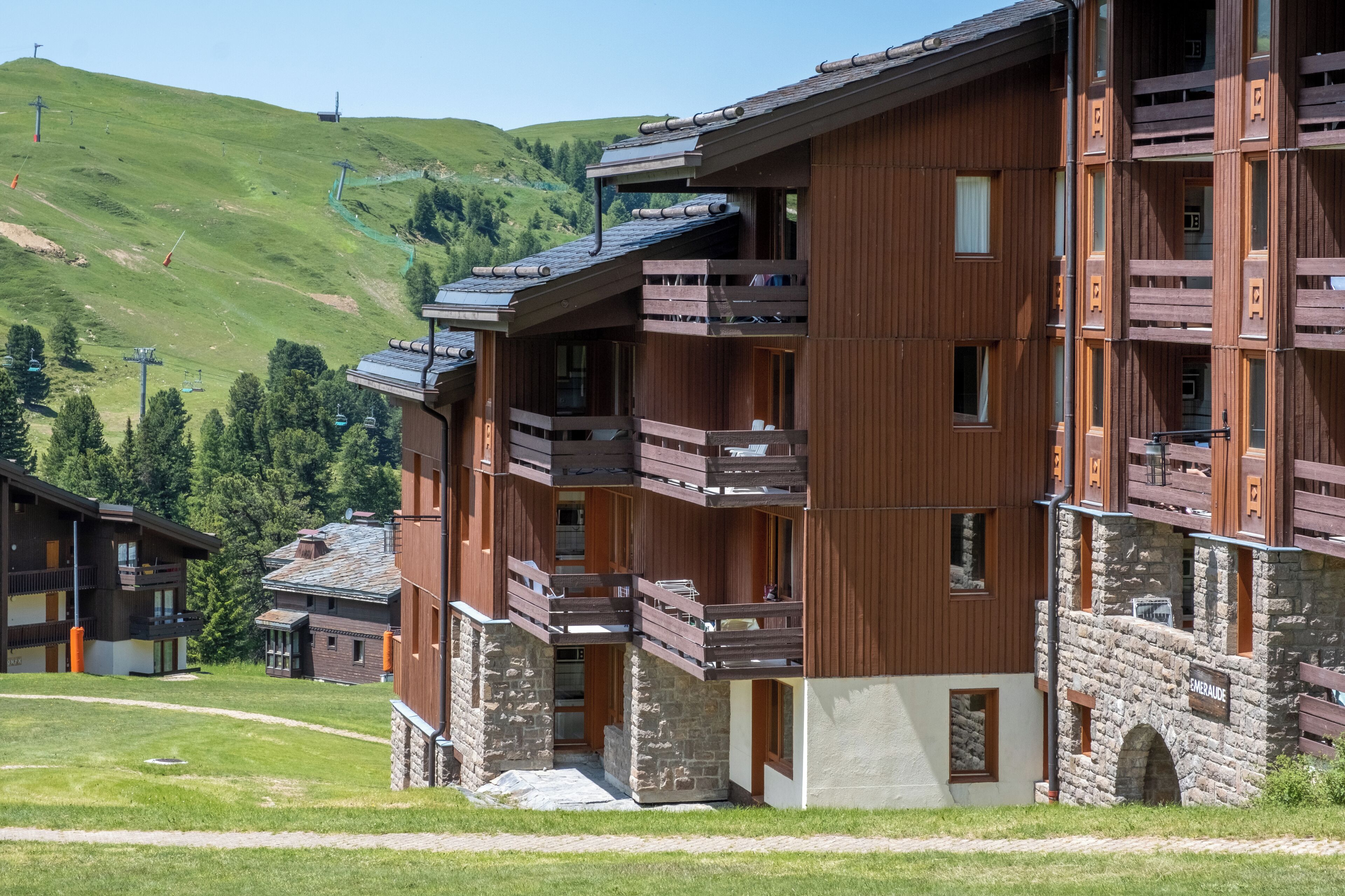 Un immeuble en bois à plusieurs étages, doté de balcons et d’une base en pierre, avec un télésiège visible en arrière-plan.