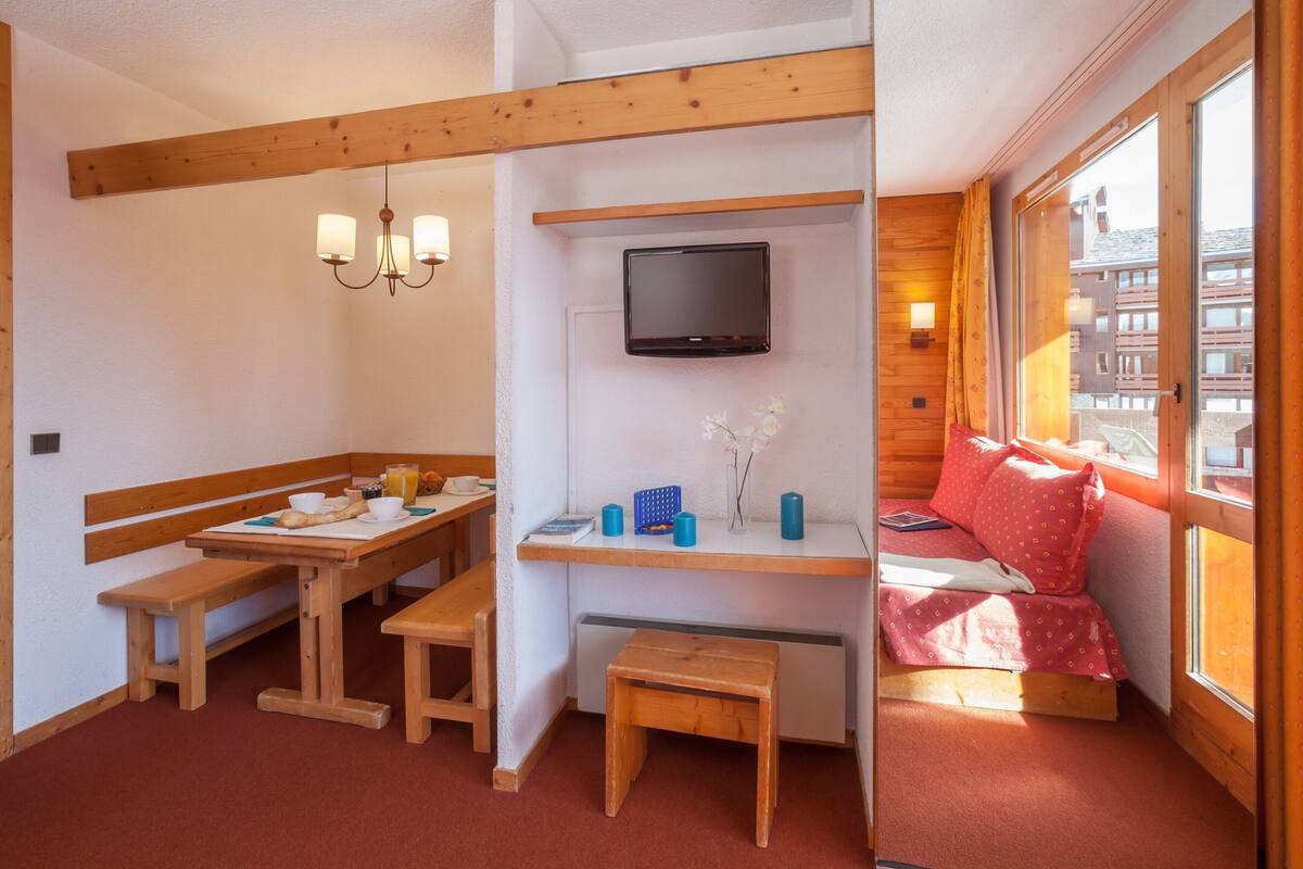 Une pièce chaleureuse avec un banc en bois, une petite table sur laquelle sont disposés des éléments pour le petit-déjeuner, un téléviseur fixé au mur et une fenêtre avec des rideaux.