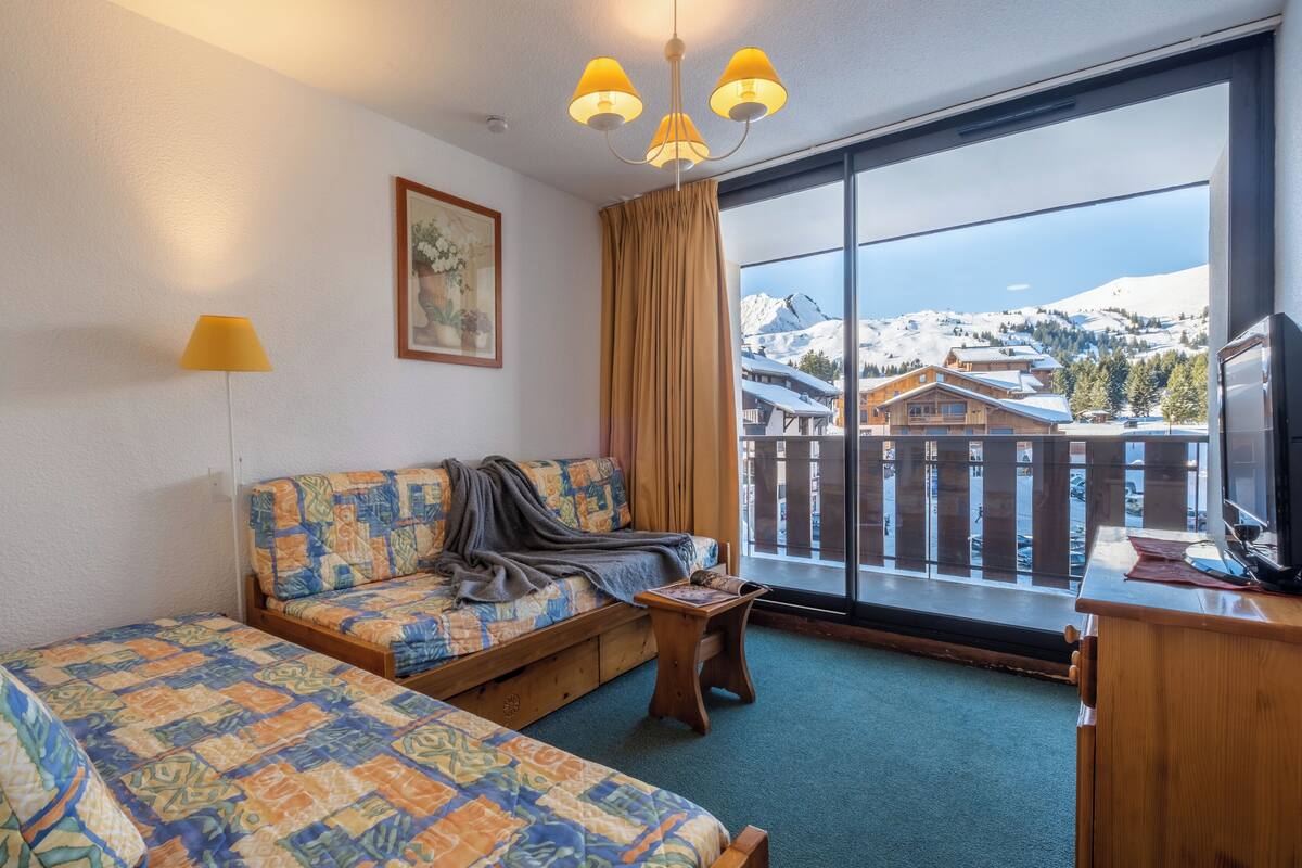 A hotel room with a large window, a sofa, two single beds, a wooden desk, and a chandelier.