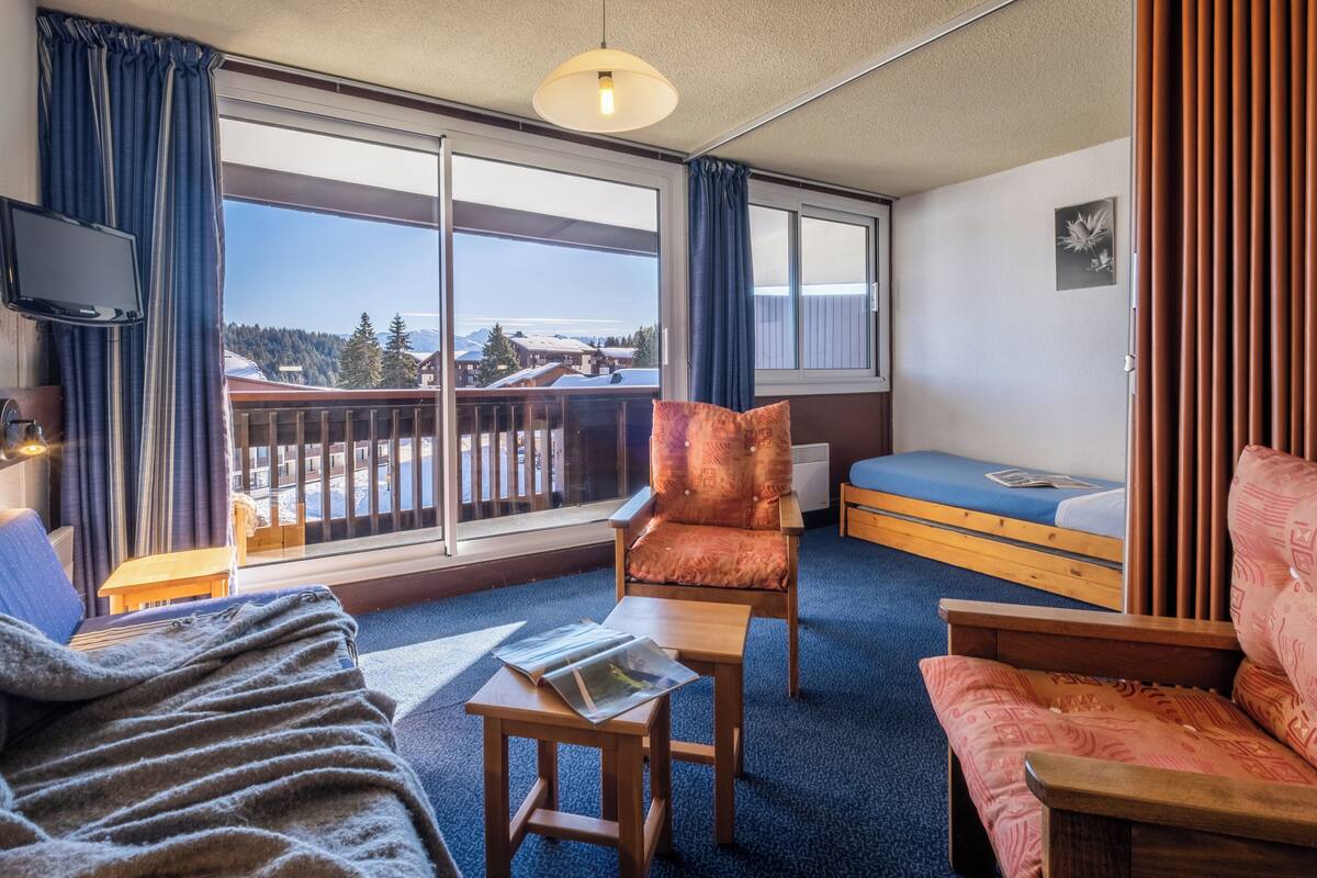 A room with a bed, a sofa, a chair, a small table, a TV, and a balcony with a view of the mountains.