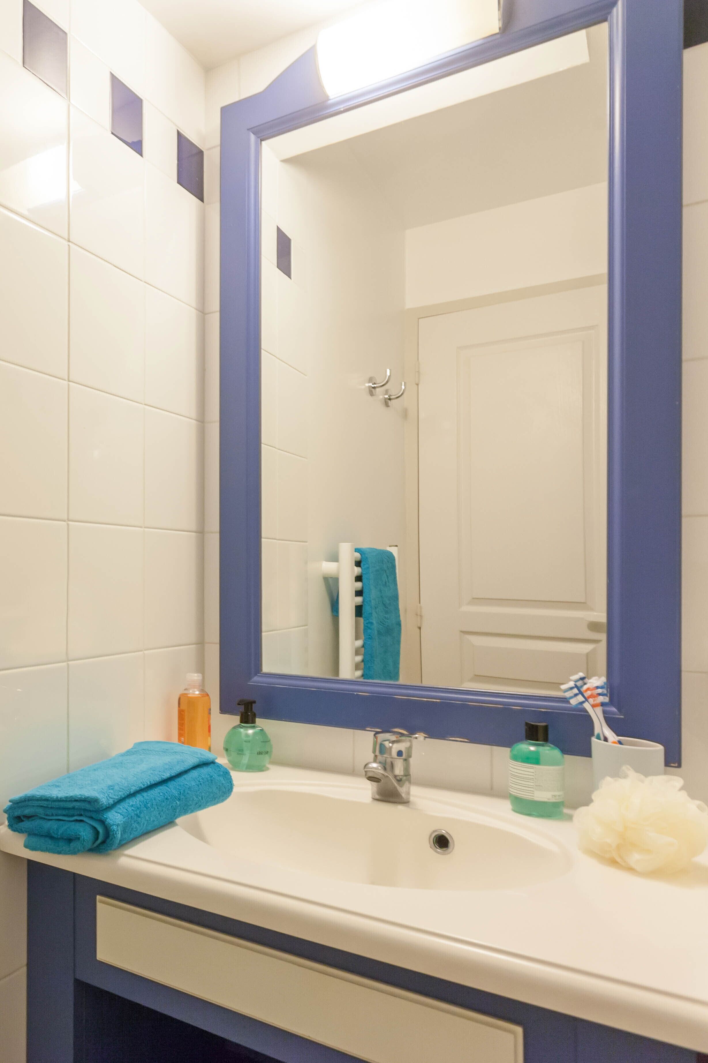 Une salle de bain avec un lavabo blanc, des murs recouverts de carreaux bleus, un grand miroir et une serviette posée sur le comptoir.