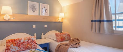 A hotel room with two beds, a nightstand, a window with curtains, and wall-mounted lamps.