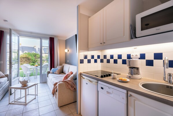 A modern kitchen with white cabinets, a microwave, and a coffee maker. There is a dining area with a table and chairs, and a sliding glass door leading to a balcony with a view of houses.