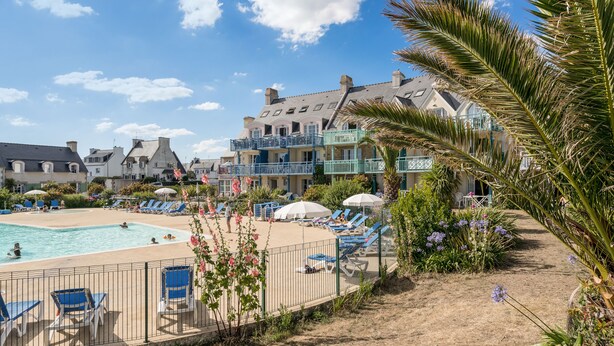 A pool area with lounge chairs and umbrellas, surrounded by a fence and palm trees. - Résidence Pierre & Vacances Cap Marine (Guilvinec)