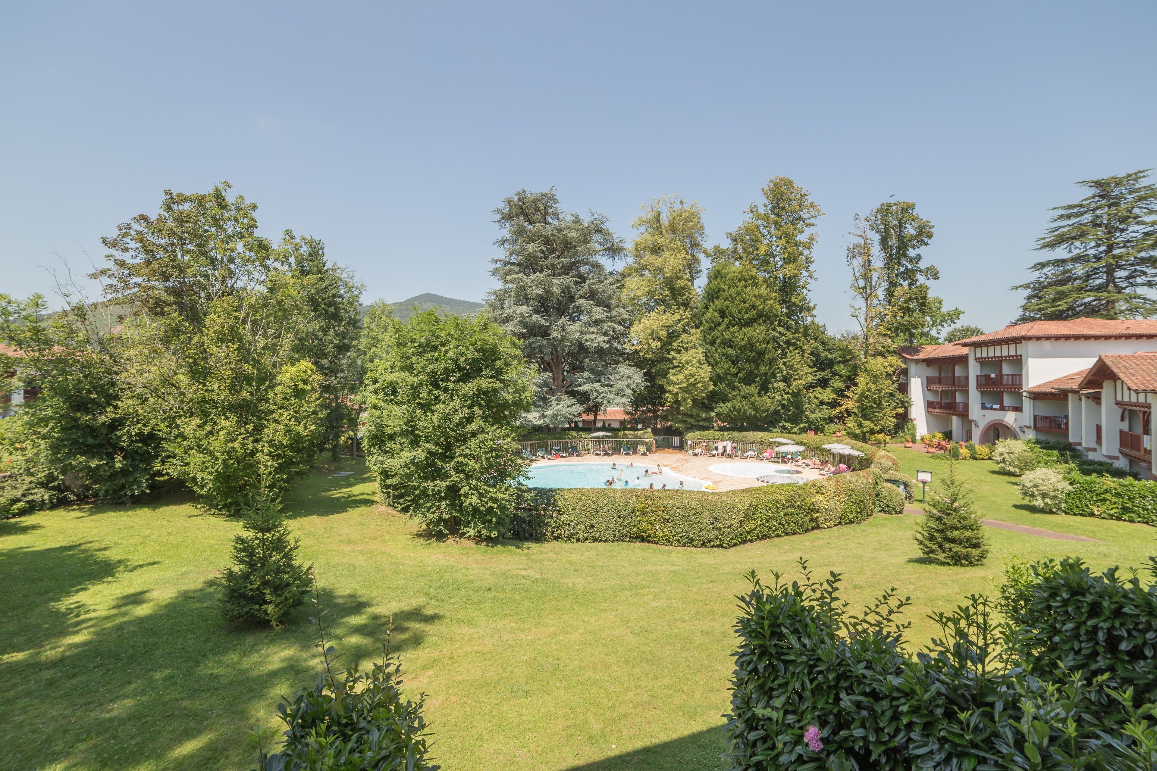 A pool surrounded by lush greenery, a well-maintained lawn, and a building with a red-tiled roof.