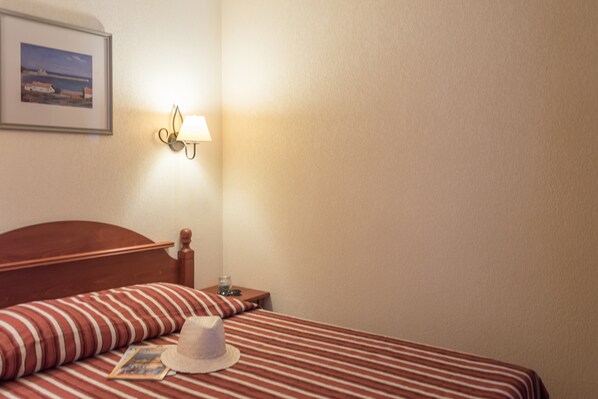 A hotel room with a wooden headboard, a striped bedspread, a white hat, a framed picture on the wall, and a bedside lamp.