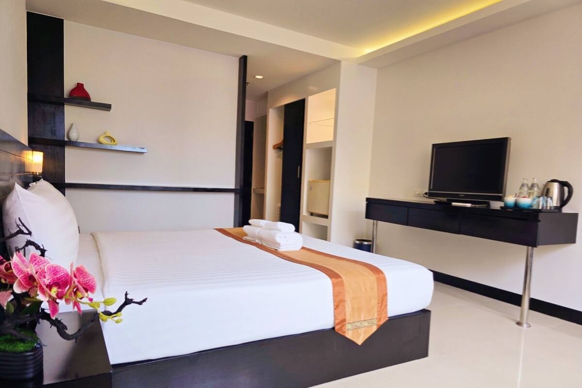 A modern hotel room with a large bed, a flat-screen TV, a desk with a coffee maker, and a potted plant.