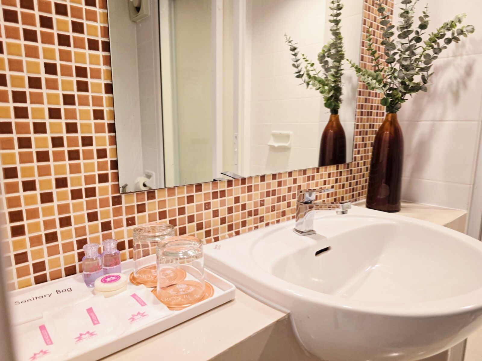 A bathroom with a white sink, a mirror, and a tiled backsplash. There are two brown vases with green plants on the wall.