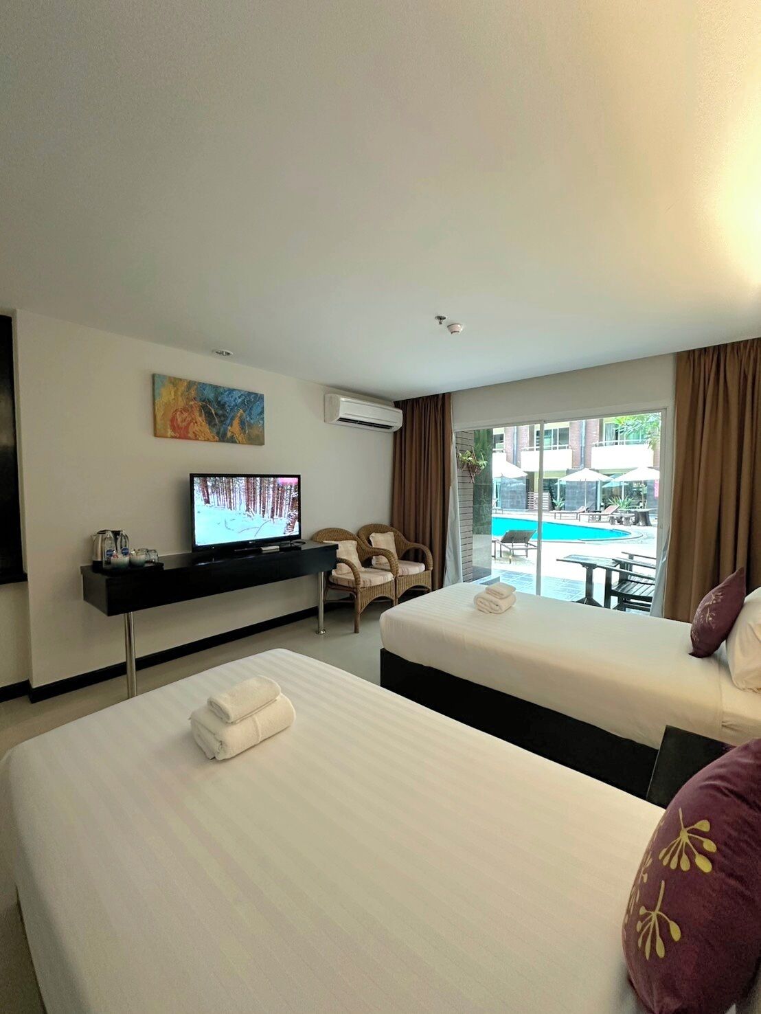 A hotel room with two beds, a TV, a desk, and a view of a pool.