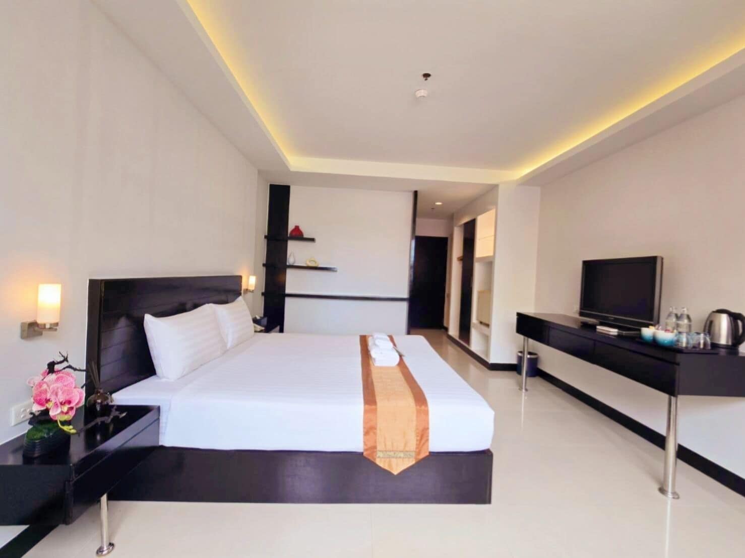 A modern hotel room with a large bed, a desk with a television, and a small table with a vase of flowers.