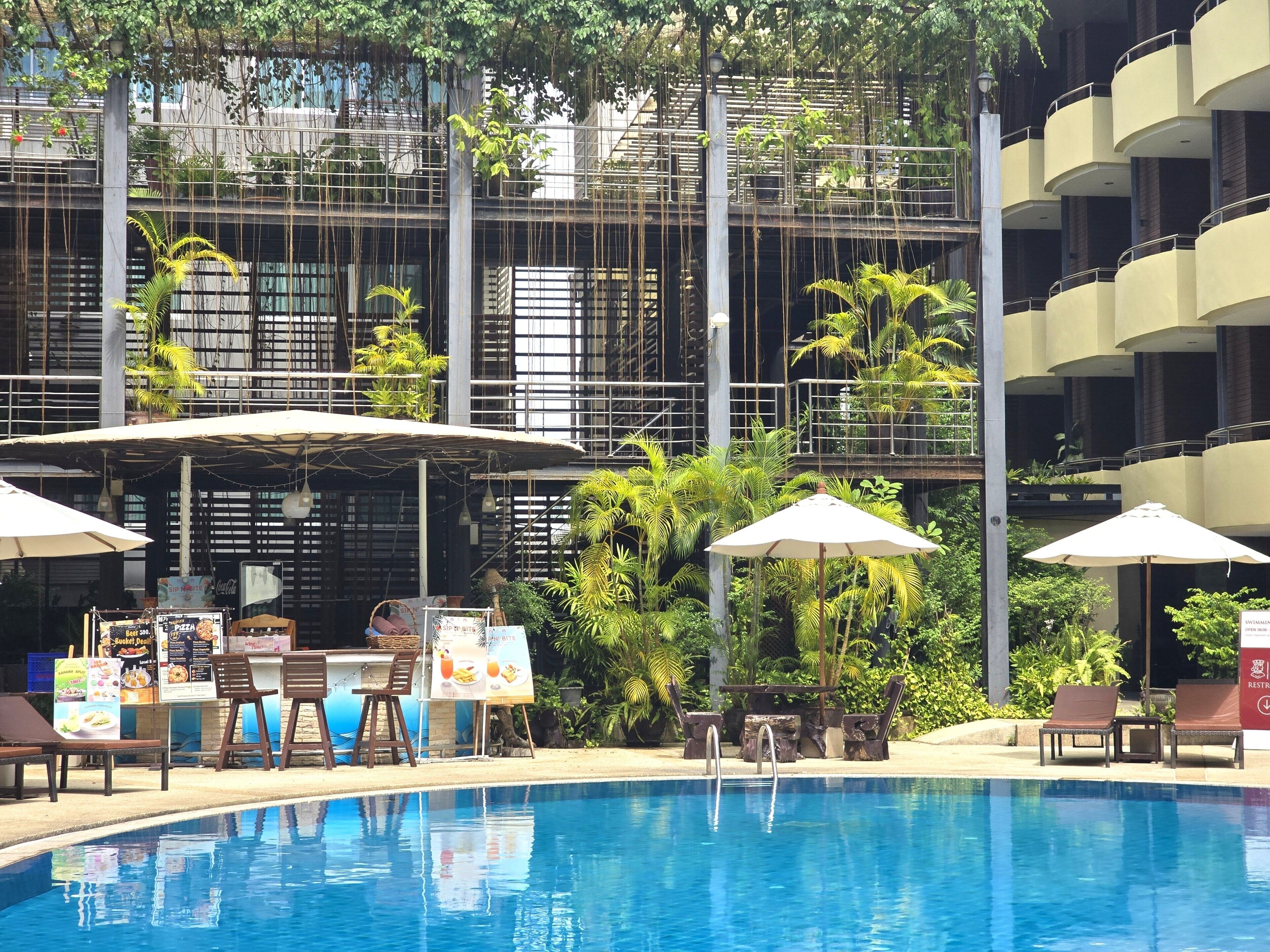 A swimming pool with lounge chairs and umbrellas, surrounded by lush greenery and a multi-story building with balconies.