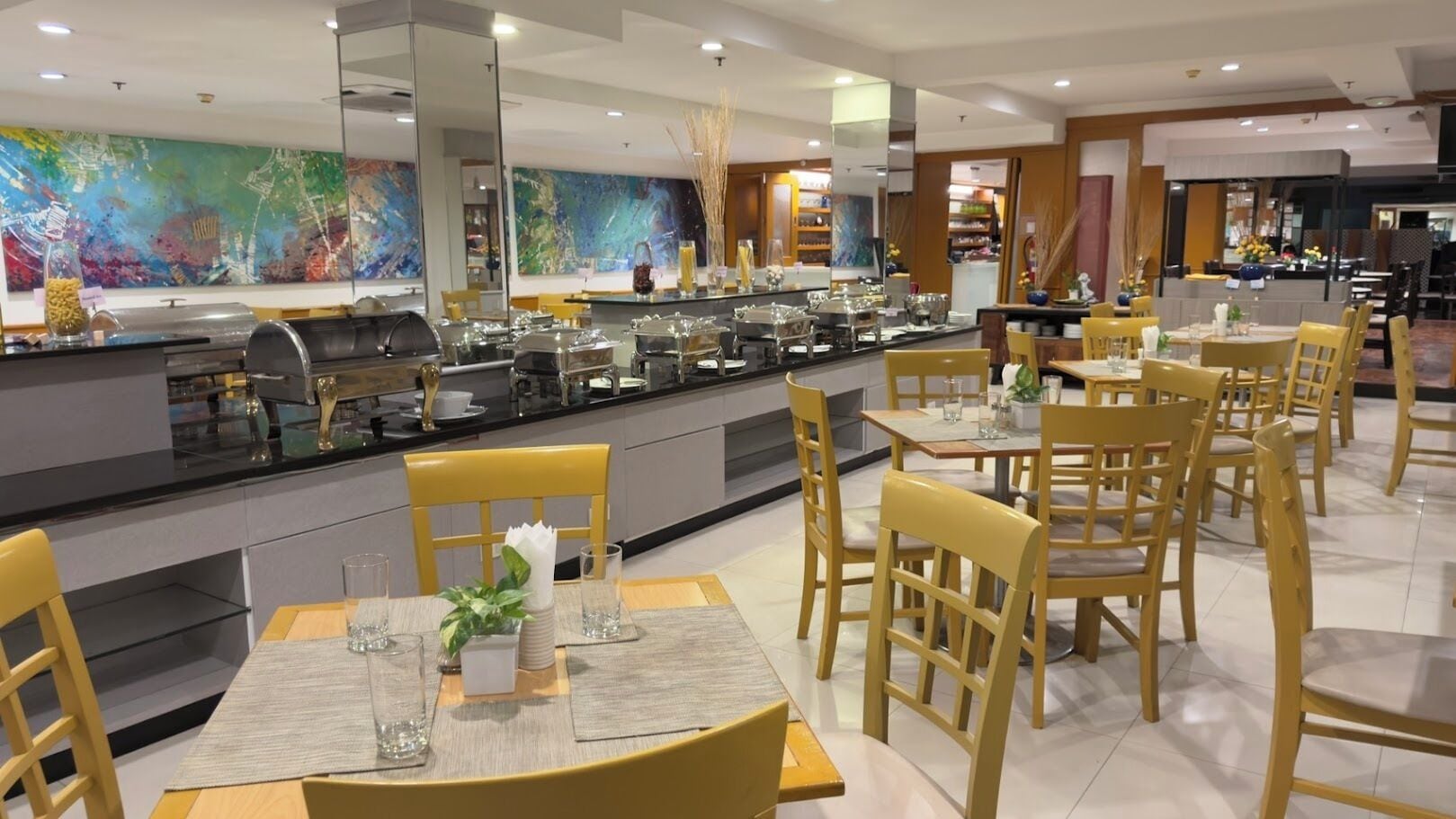 A modern restaurant with yellow chairs and tables, a buffet counter, and abstract art on the walls.