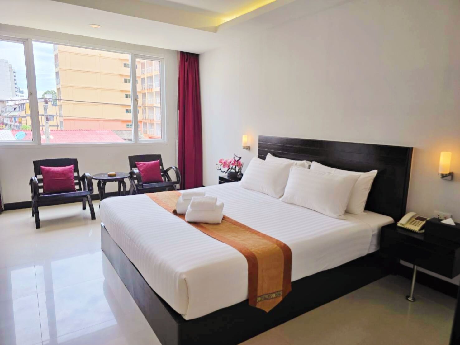 A hotel room with a large bed, a bedside table with a lamp, a chair, and a view of buildings through a window.