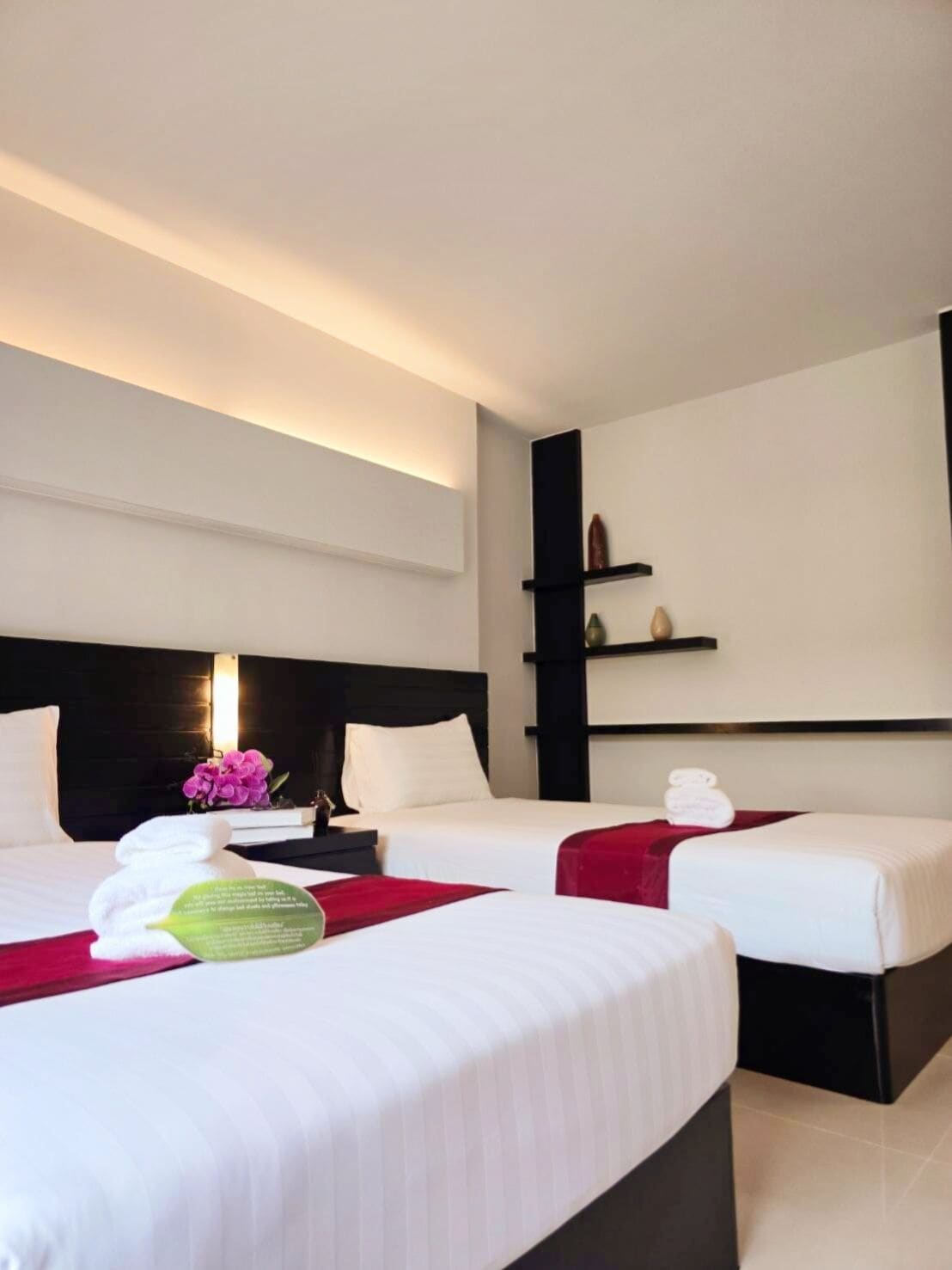 A hotel room with two beds, a black headboard, a bedside table with a lamp, and a shelf with decorative items.