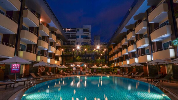 A swimming pool with lounge chairs and umbrellas, surrounded by multi-story buildings with balconies. - Baron Beach Hotel (Pattaya)