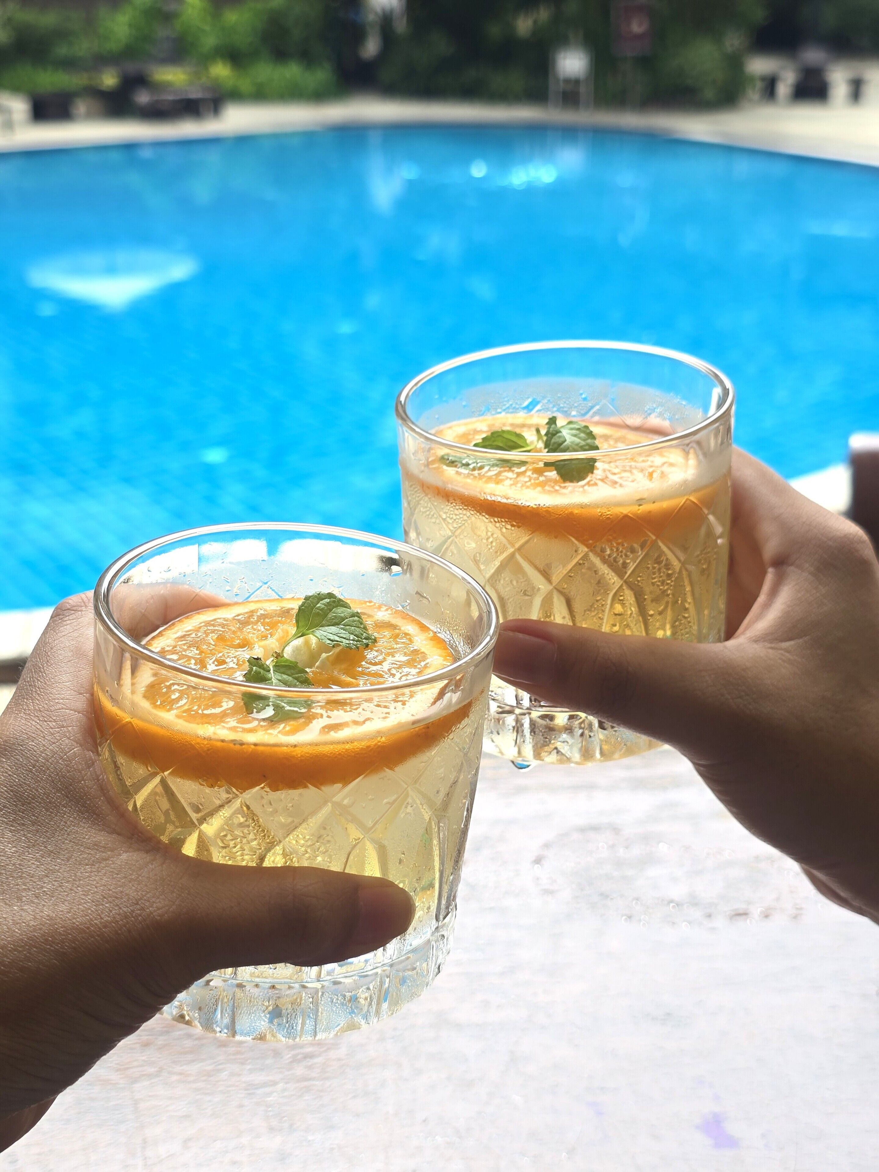 Due bicchieri di bevanda con guarnizione, tenuti da mani, di fronte a una piscina.