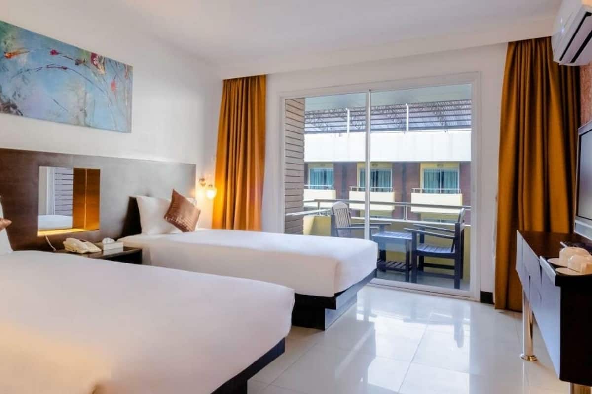 A hotel room with two beds, a TV, a desk, and a balcony with a view of buildings.