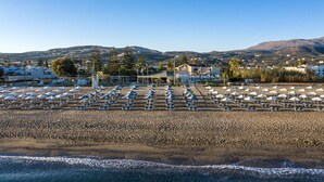 A beach with rows of umbrellas and lounge chairs, a building with a red roof, and mountains in the background.