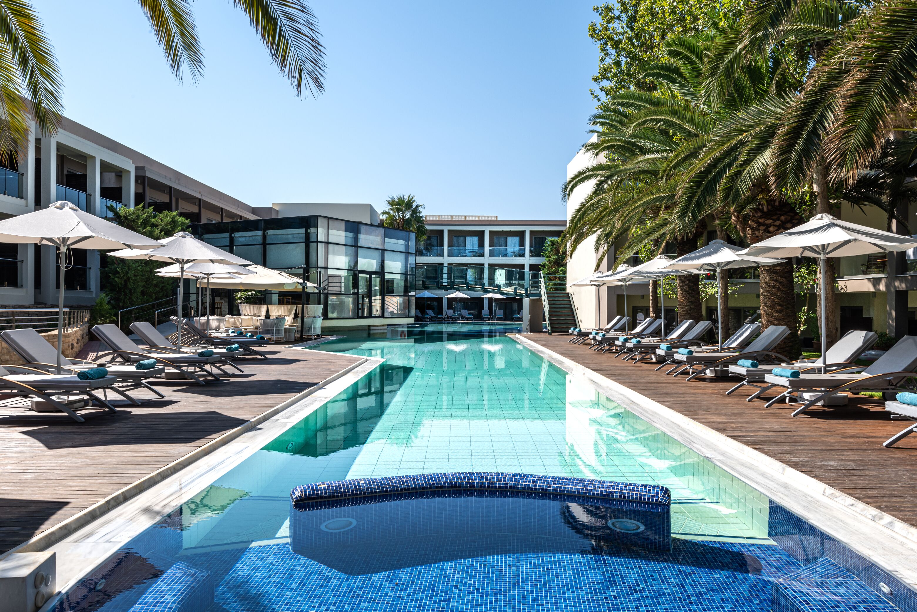 A swimming pool with lounge chairs and umbrellas, surrounded by palm trees and modern buildings.