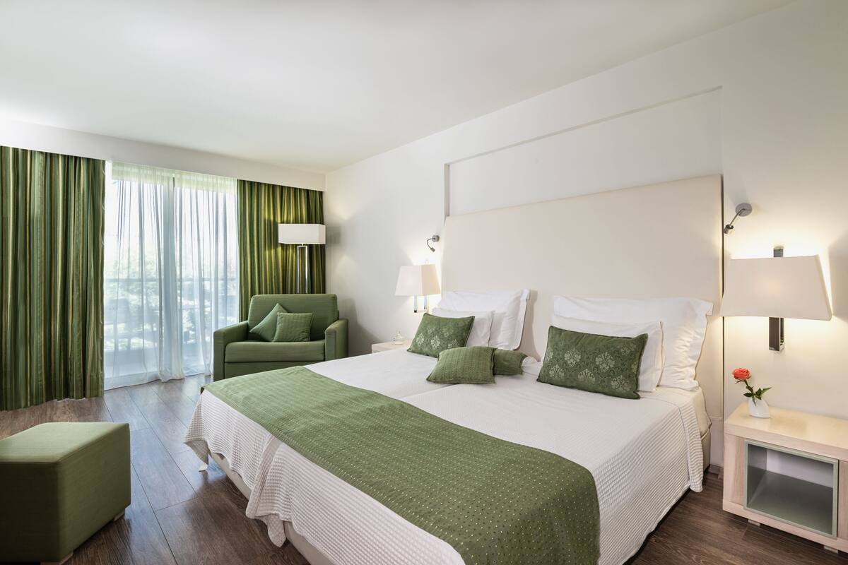 A hotel room with a large bed, a green armchair, a nightstand with a lamp, and a window with curtains.