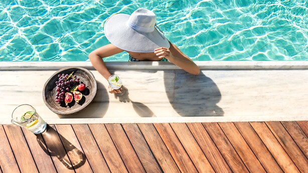 A person relaxing by a pool with a bowl of fruit and a drink on the deck.