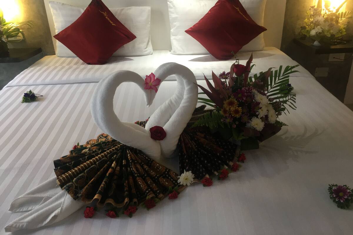 Una cama bien hecha con una decoración de cisne en forma de corazón, almohadones rojos y un arreglo floral.