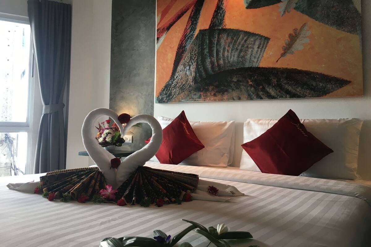 Habitación de hotel con una cama, un centro de mesa con forma de corazón, almohadones rojos y un cuadro abstracto grande en la pared.