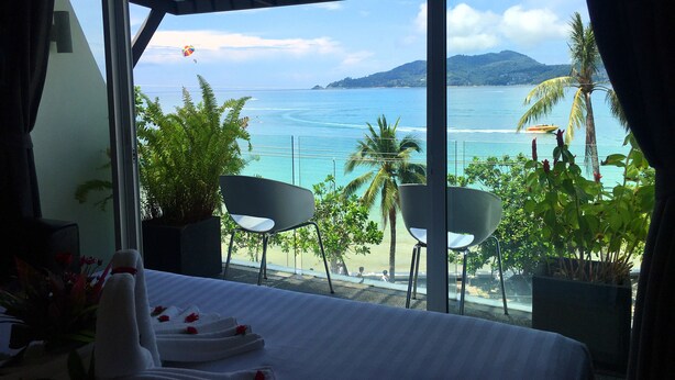 A hotel room with a large window overlooking a beach and ocean. There is a bed with white linens and a small table with a vase of flowers. - The Front Hotel and Apartment (Patong)
