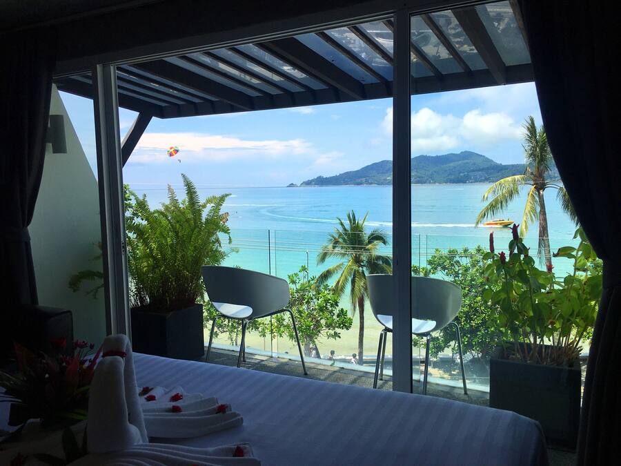 Habitación de hotel con un ventanal grande que da a una playa y al océano. Hay una cama con ropa de cama blanca y una mesita con un florero.