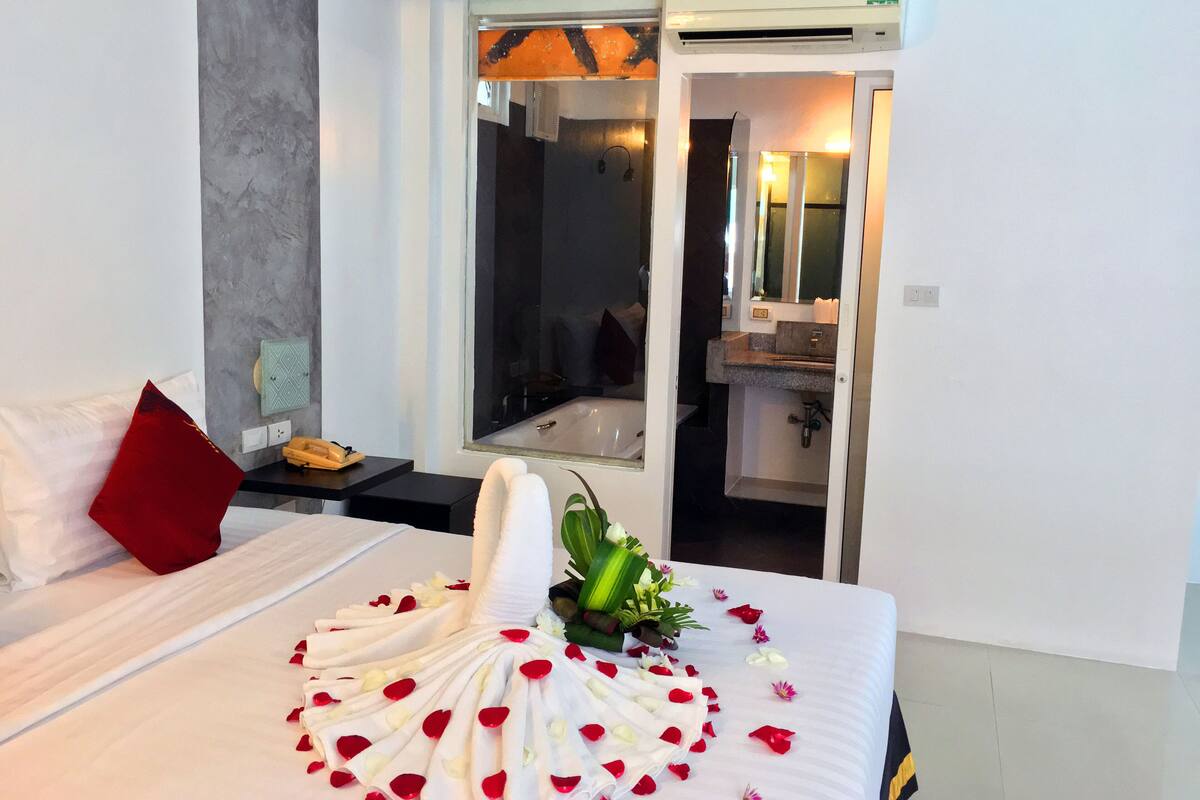 Una cama bien hecha con una toalla y pétalos de flores, un cojín rojo, una mesita de noche con un teléfono y una ventana con aire acondicionado.