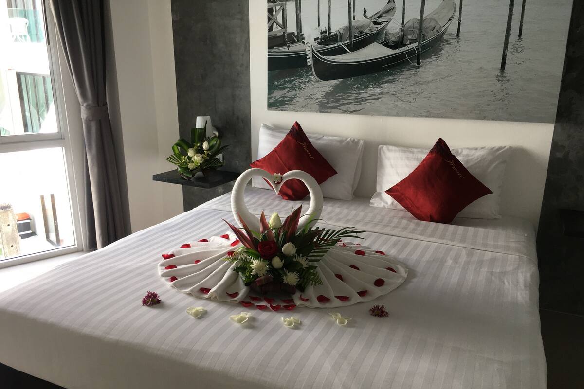 Una cama bien tendida con un centro de mesa con forma de cisne, almohadas rojas y un cuadro con barcos en la pared.