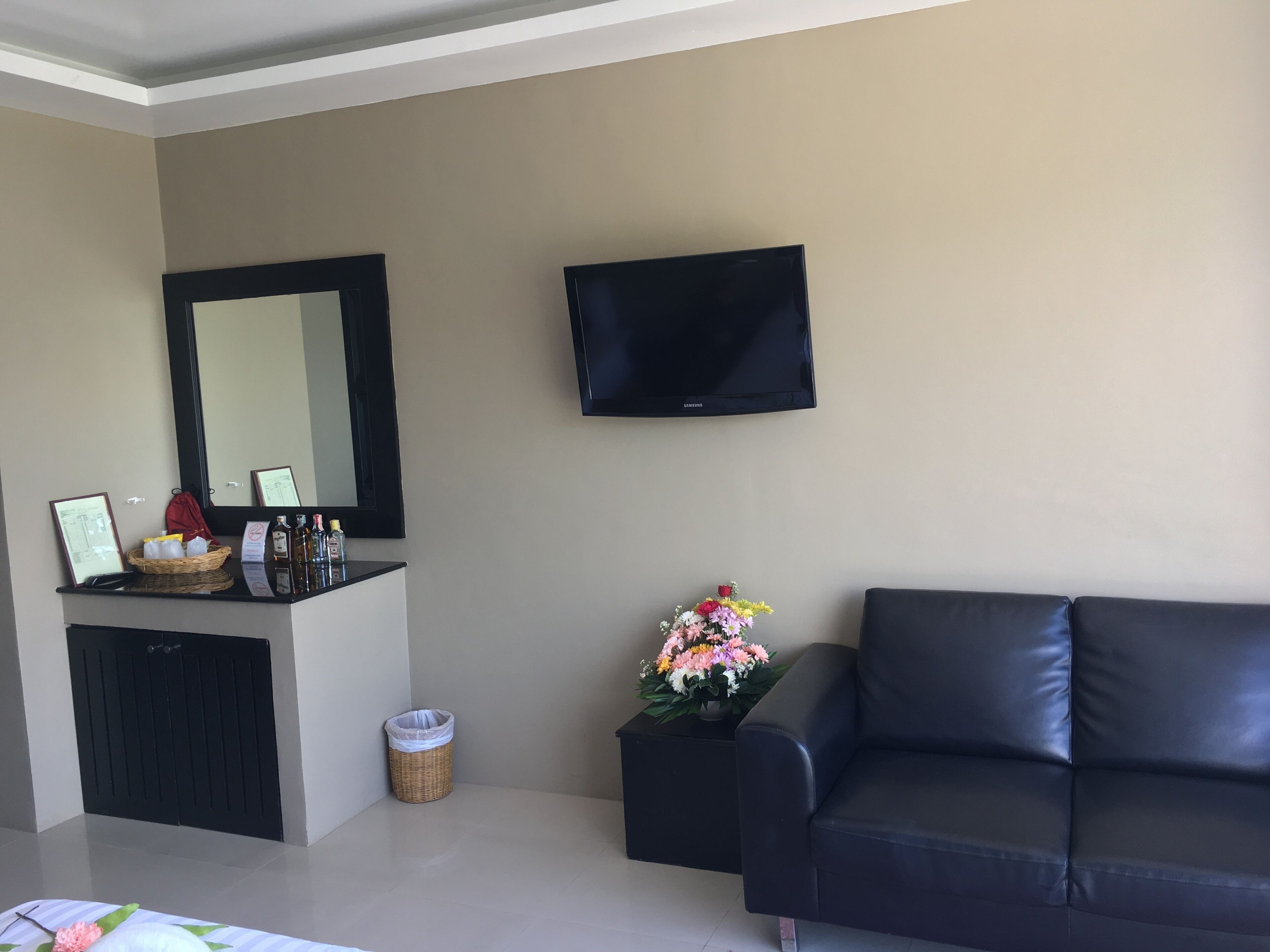 Een woonkamer met een zwarte bank, een flatscreen-tv, een grote spiegel, een klein tafeltje met een mand en flessen, en een prullenbak.