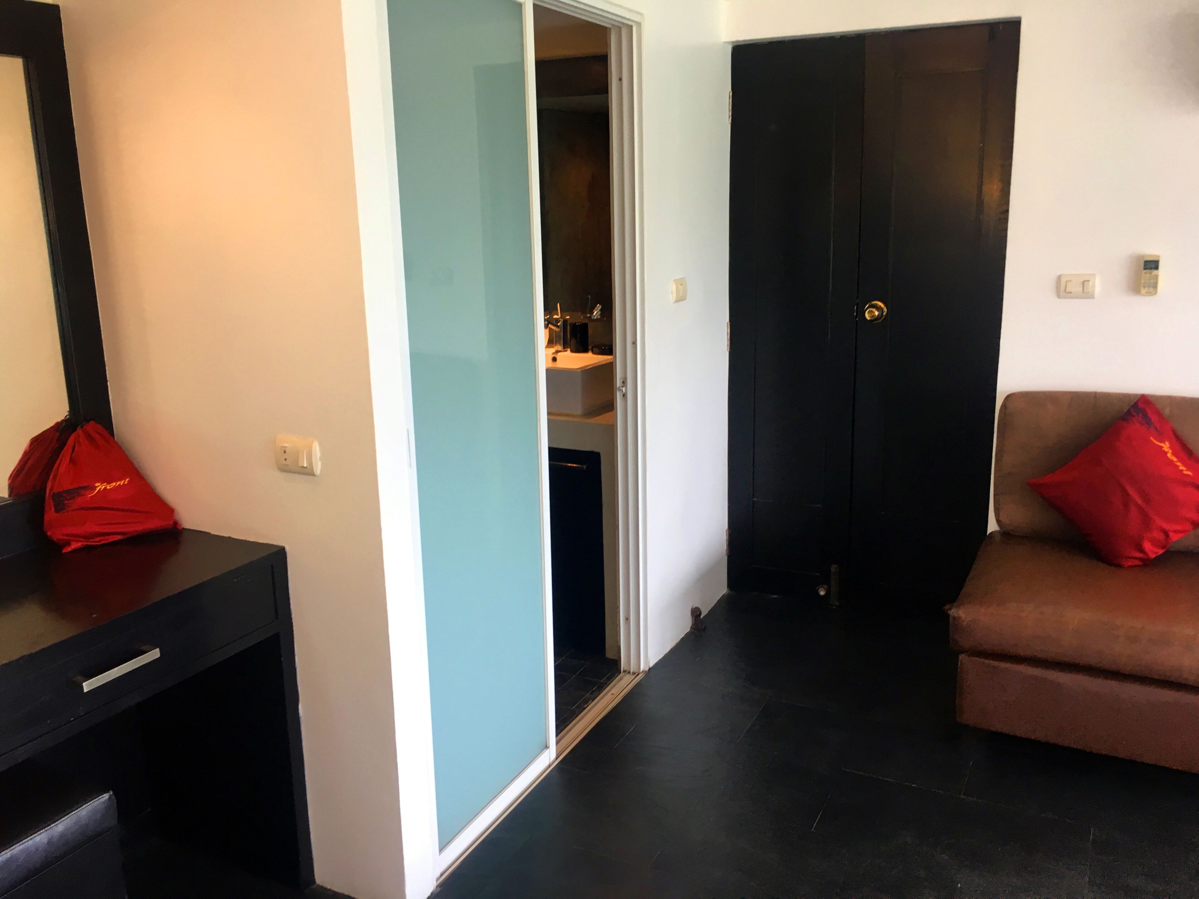 Ein Zimmer mit einem schwarzen Schreibtisch, einer roten Tasche, einem braunen Sofa mit rotem Kissen und einer Tür zu einem Badezimmer.