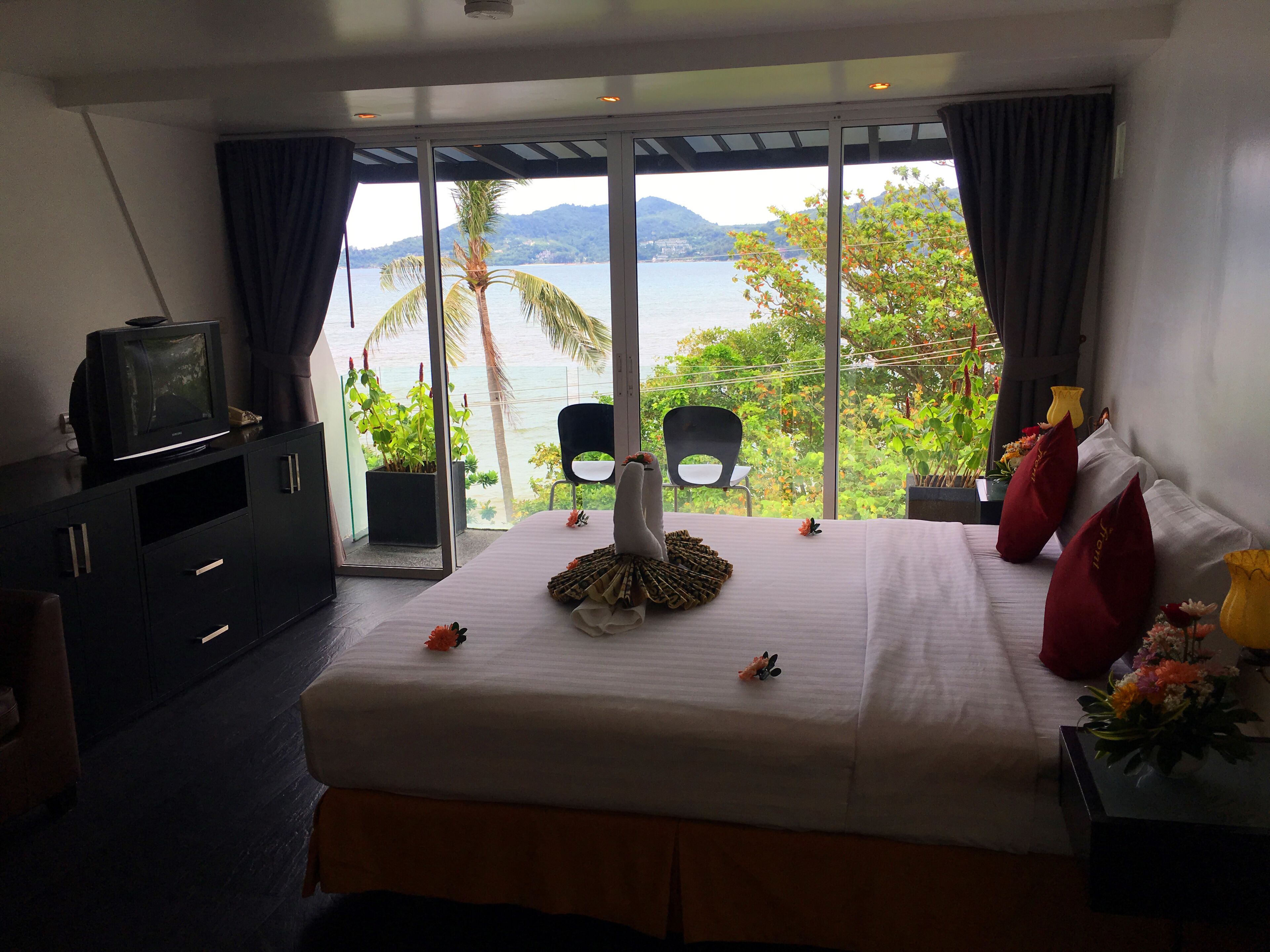 A hotel room with a bed, a TV, a chair, and a view of the sea and mountains.