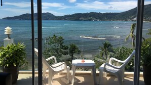 A balcony with two white plastic chairs and a small table, overlooking a coastal view with clear blue water and distant hills. - The Front Hotel and Apartment (Patong)