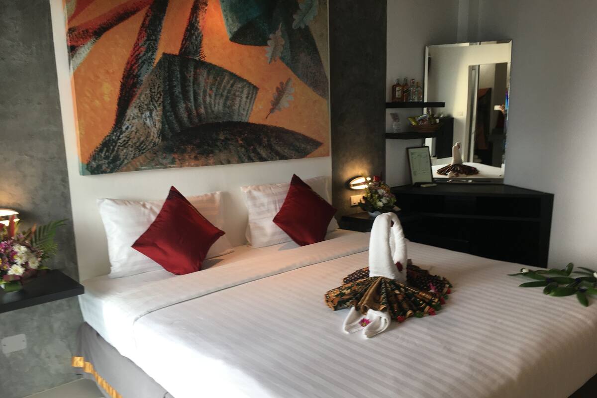 Habitación de hotel con una cama grande, un cuadro en la pared, un espejo y una mesita de noche con una lámpara.