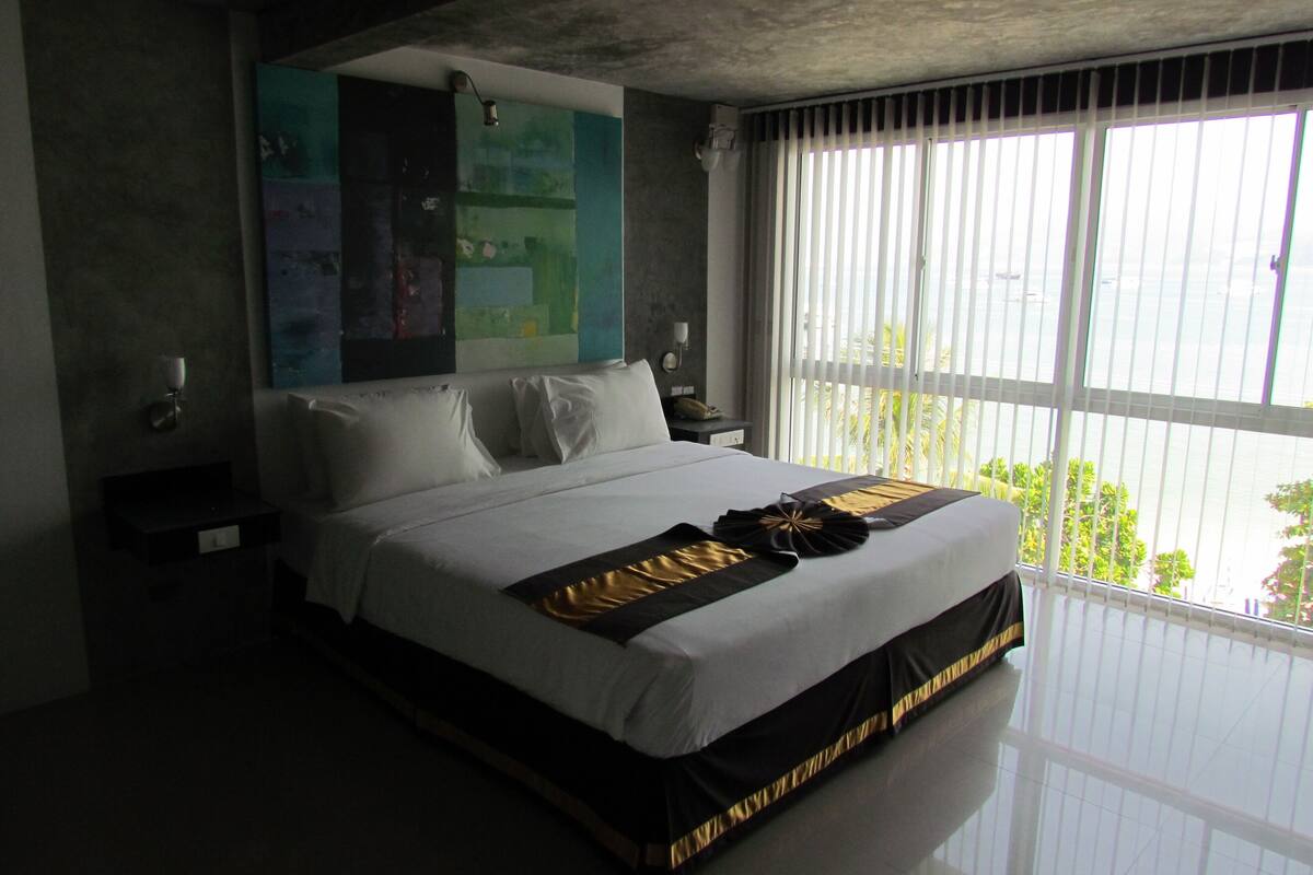 Habitación de hotel moderna con una cama grande, una mesita de noche, una lámpara de pared y un ventanal con persianas.