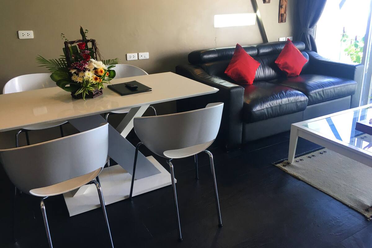 Una sala de estar moderna con un sofá de cuero negro, sillas blancas, una mesa blanca y un jarrón con flores.
