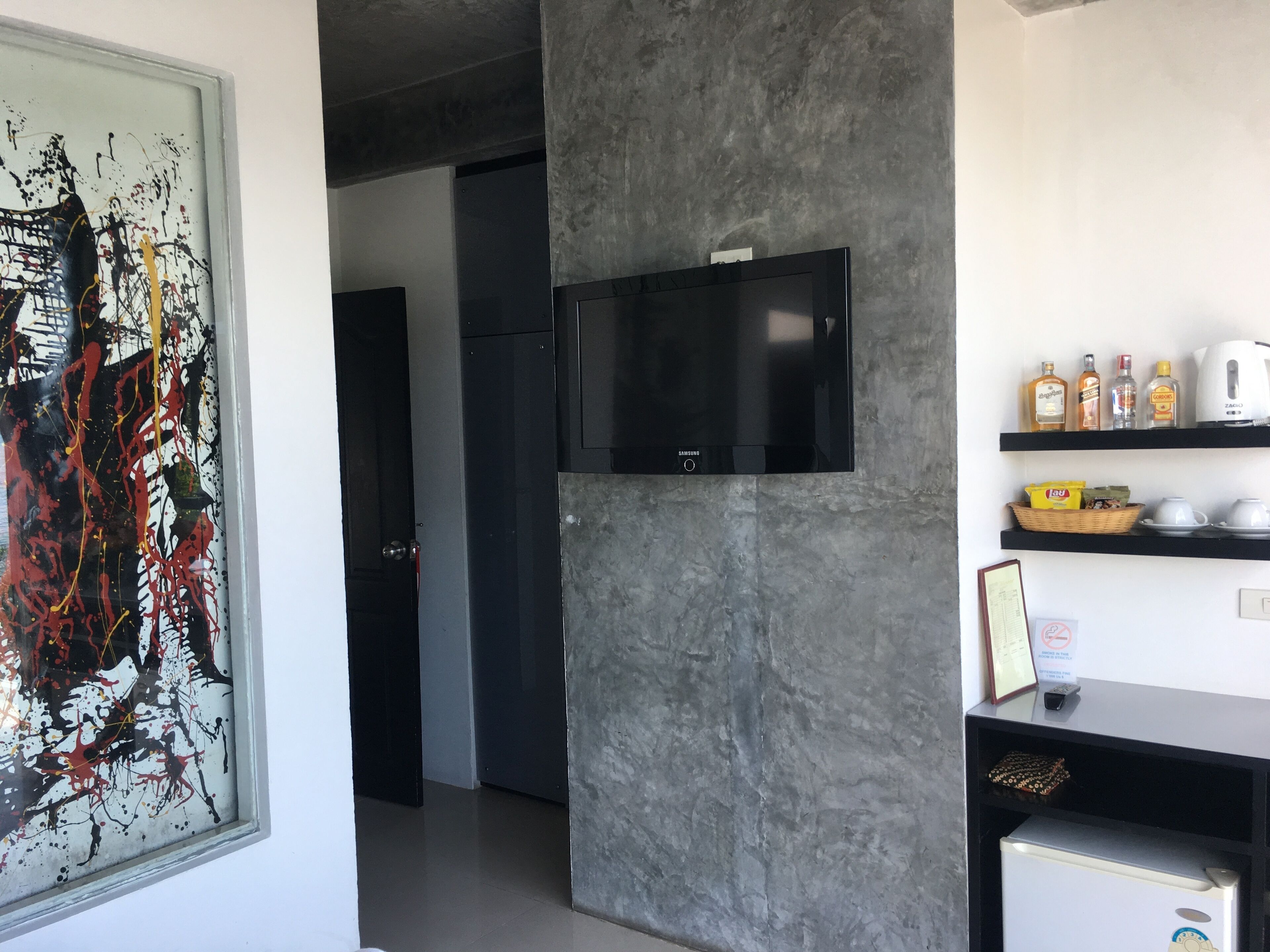 A modern kitchen with a flat-screen TV mounted on a grey wall, a refrigerator, and a shelf with kitchen items.