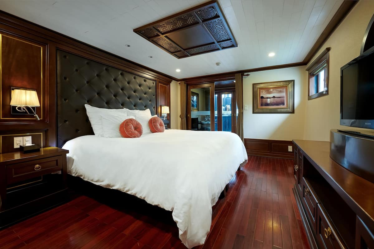 A large, neatly made bed with a tufted headboard, a bedside table with a lamp, and a flat-screen TV on a stand.