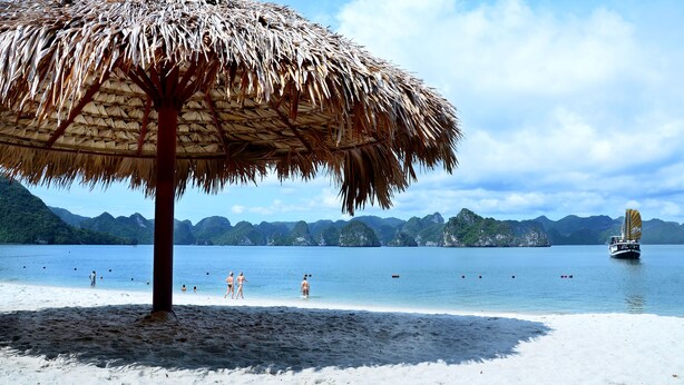 A beach with a thatched umbrella, clear blue water, and distant mountains. - Paradise Peak Cruise (Tuan Chau)