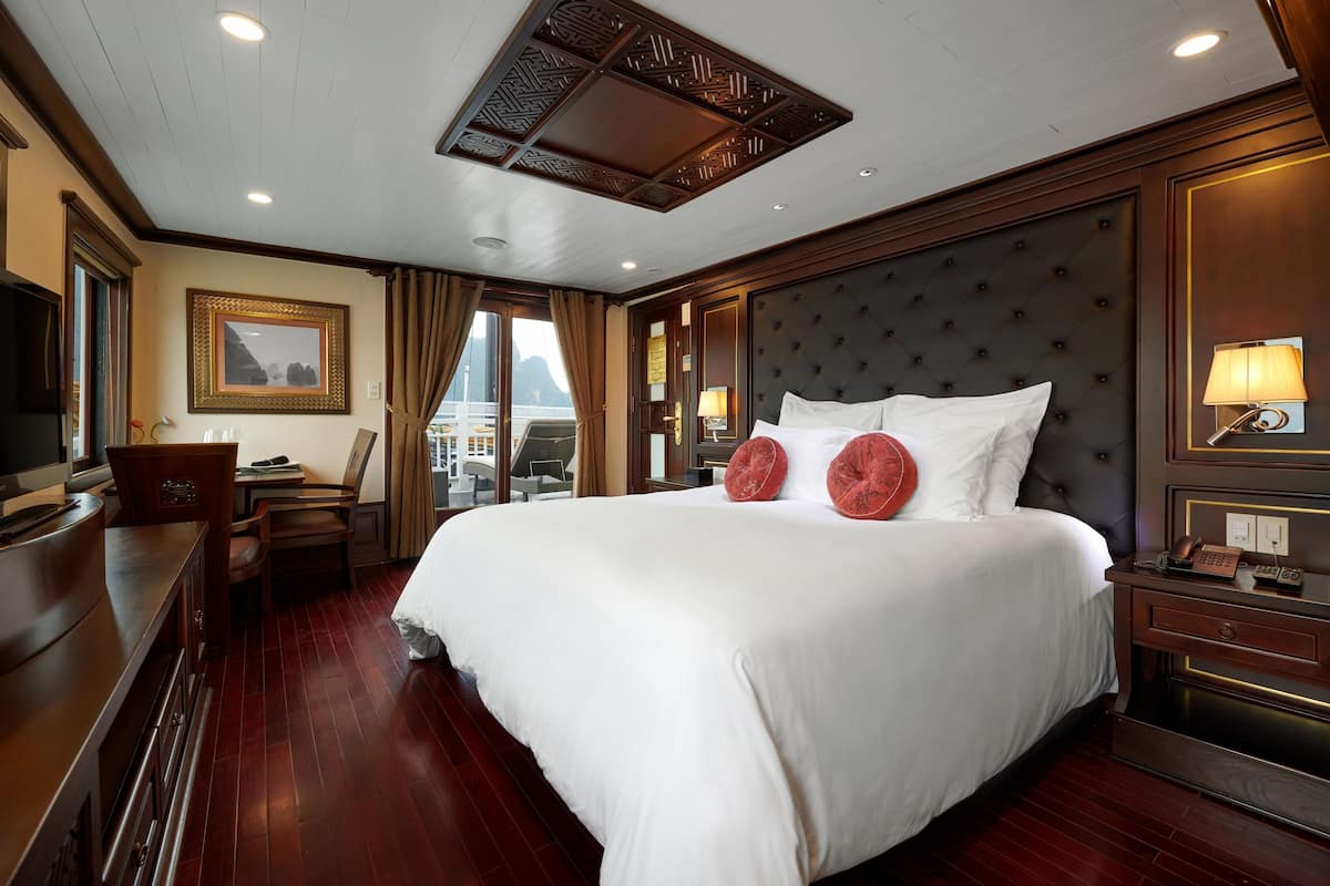 A large bed with white linens, a dark tufted headboard, and two red pillows. There is a wooden desk with a chair, a bedside table with a lamp, and a framed picture on the wall.