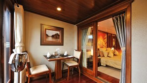 A room with a wooden floor, a dining table with chairs, a bed with yellow bedding, and a large mirror reflecting the room. - Paradise Peak Cruise (Tuan Chau)