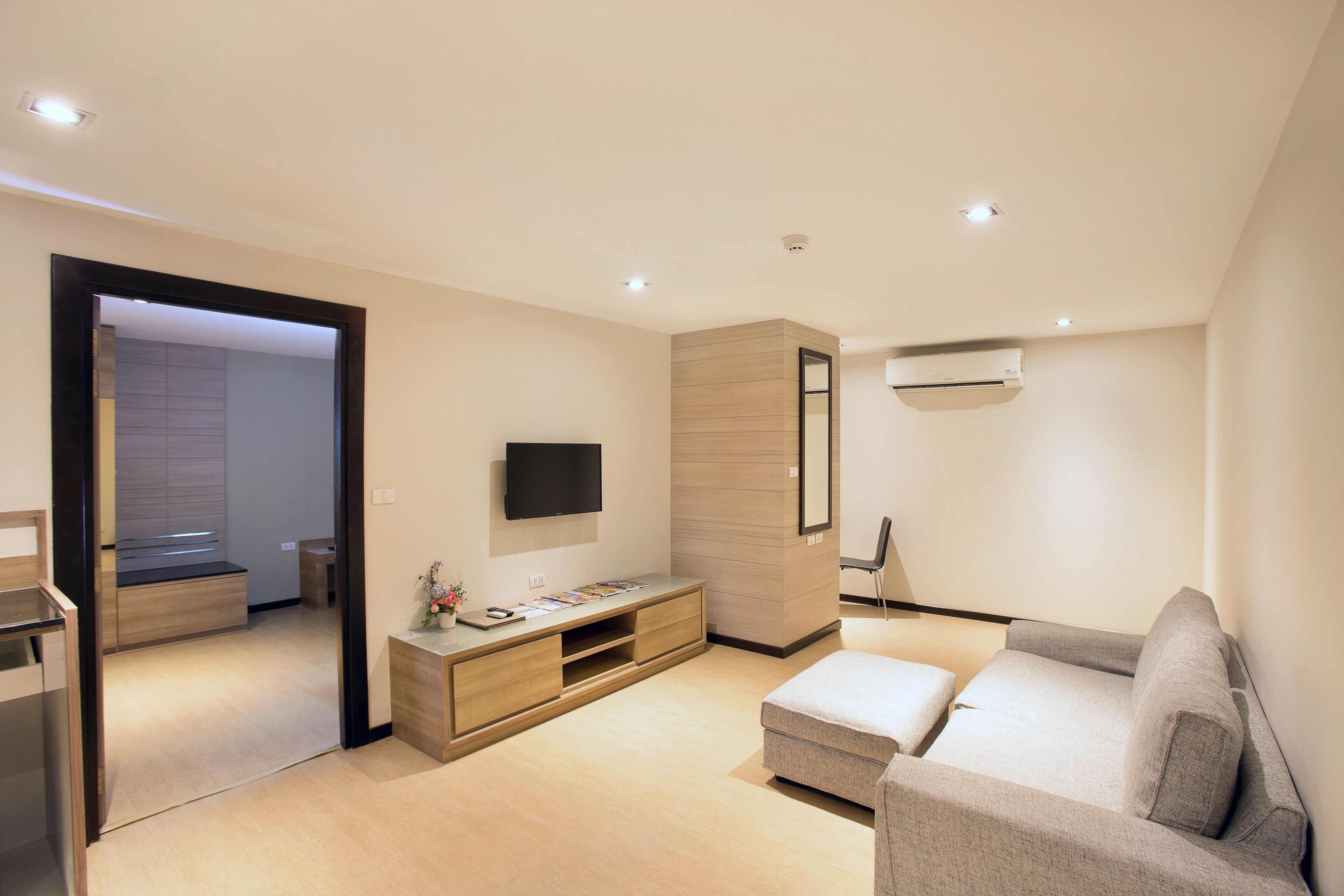 A modern living room with a flat-screen TV, a wooden entertainment unit, a grey sofa, and a small table with a vase.