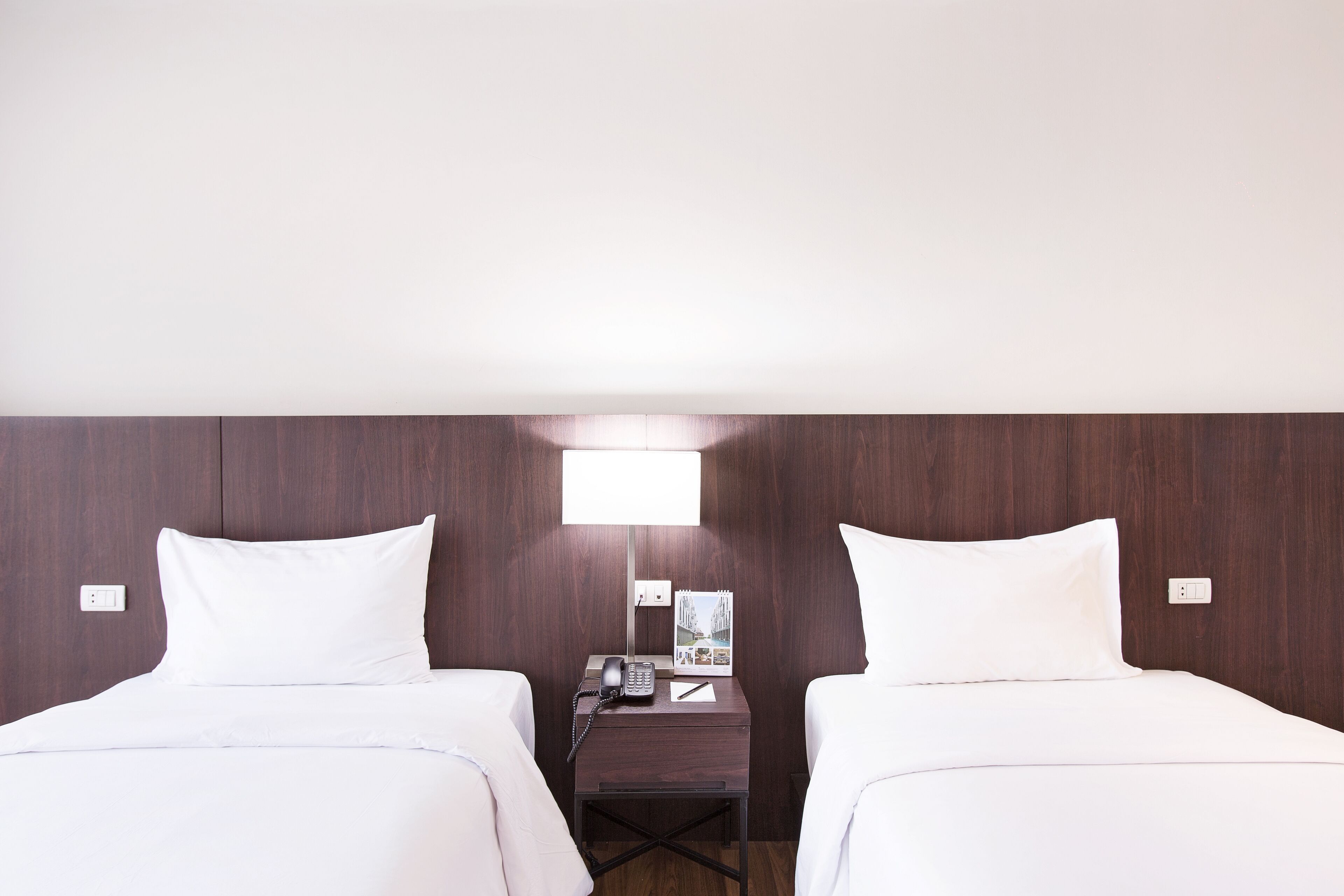 A hotel room with two single beds, a nightstand with a telephone, a lamp, and a small table with a brochure.