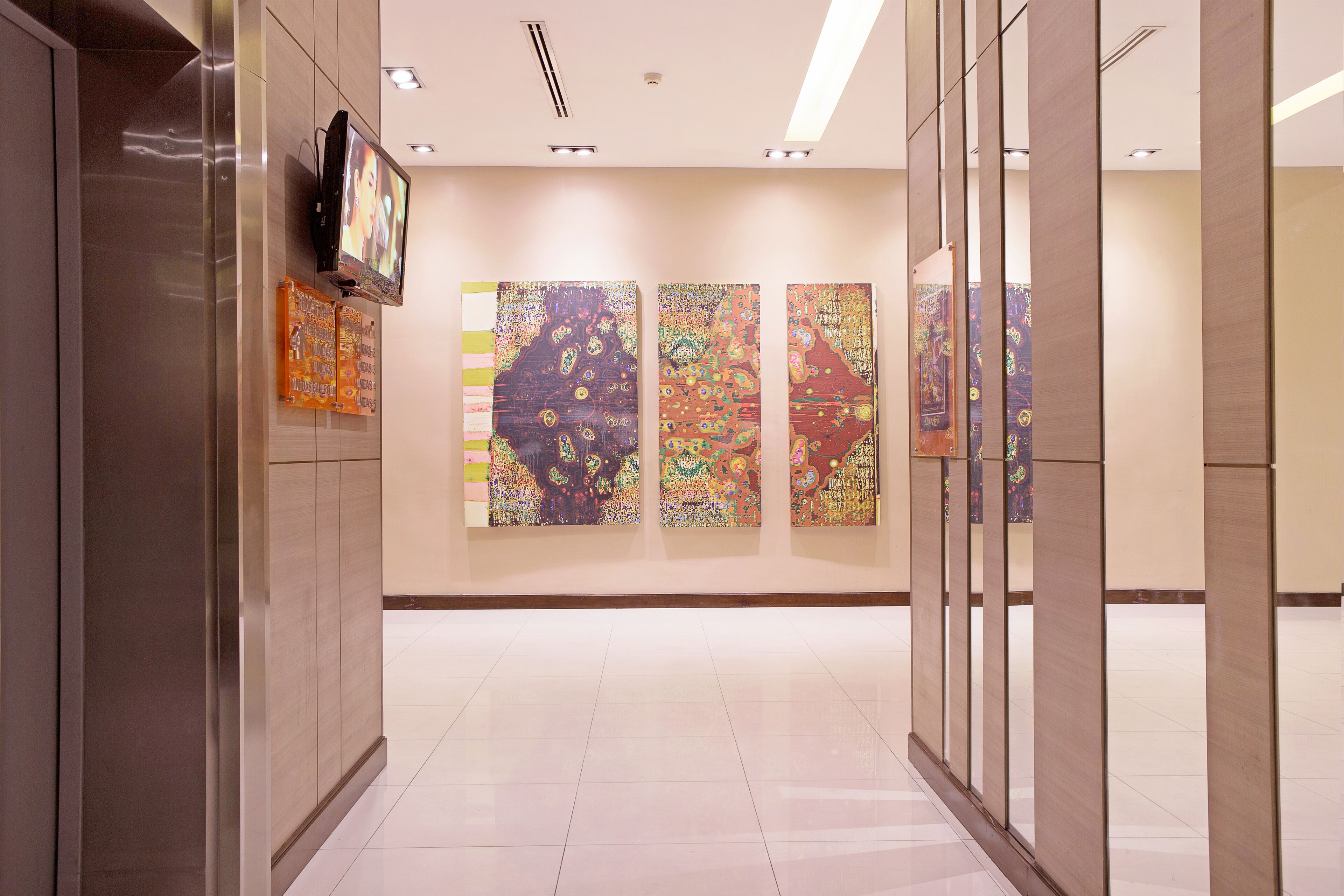A hallway with three large abstract paintings on the wall, a television mounted on the left, and a modern interior design with a tiled floor.
