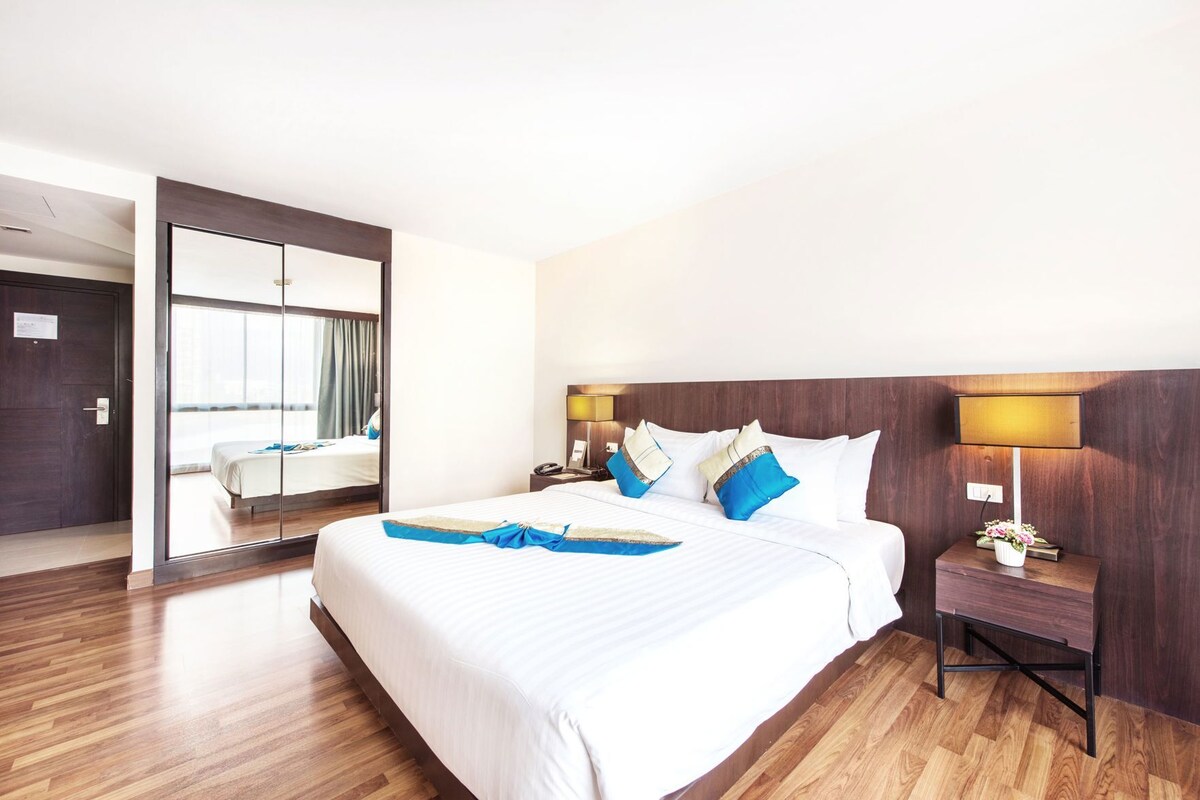 Habitación de hotel con una cama grande, suelo de madera, una mesita de noche con lámpara y vista a otra habitación a través de una puerta de vidrio.