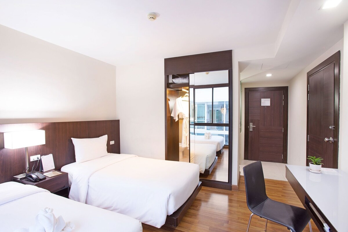 Habitación de hotel con dos camas, un escritorio, una silla, una mesita de noche, una lámpara y vistas al balcón.
