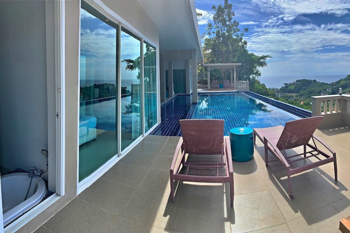 A modern balcony with a swimming pool, two wicker chairs, and a view of the surrounding landscape.